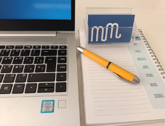 Parte del teclado de un ordenador portátil junto a una agenda y un tarjetero con el logotipo de Marta Mozo Traducciones, dos letras m minúsculas enlazadas en color blanco sobre fondo azul.