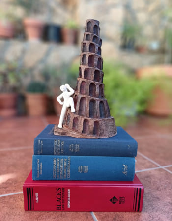 torre_babel_diccionarios Una versión esculpida en barro de la Torre de Babel, símbolo de la traducción, se sostiene sobre una pequeña pila de tres gruesos diccionarios de términos jurídico-económicos. Este grupo está colocado en primer plano sobre el suelo de un patio con un fondo difuminado de plantas en macetas.